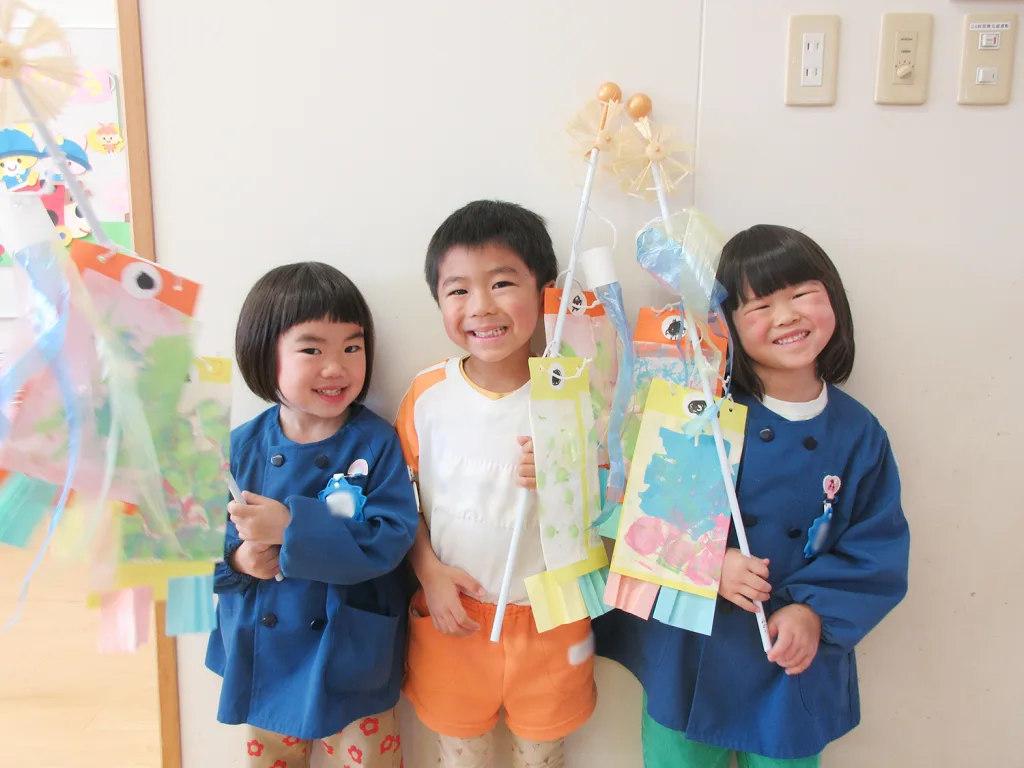 Three smiling children pose together, holding colorful paper crafts on sticks and wearing bright blue coats (center child in orange shorts).
