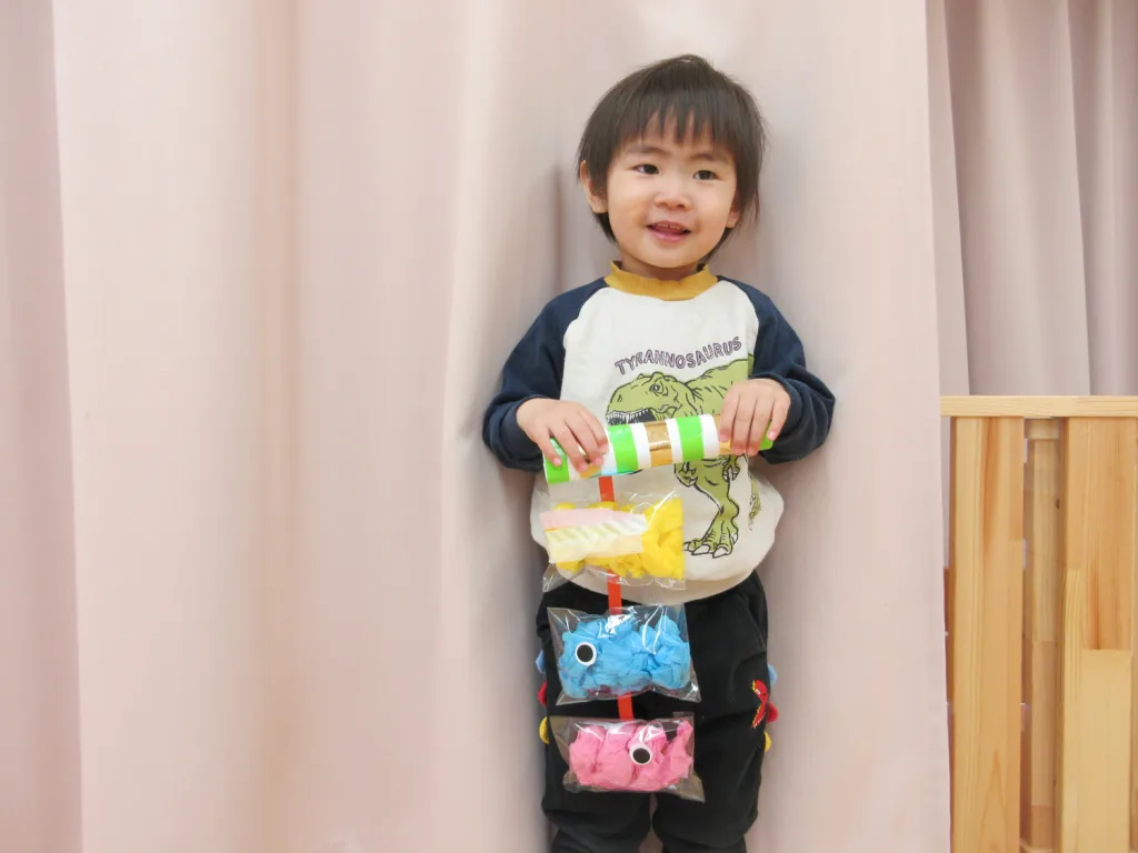 Smiling toddler holding a vertical hanging craft made of clear bags with colorful tissue shapes on a red ribbon, standing between pink curtains.