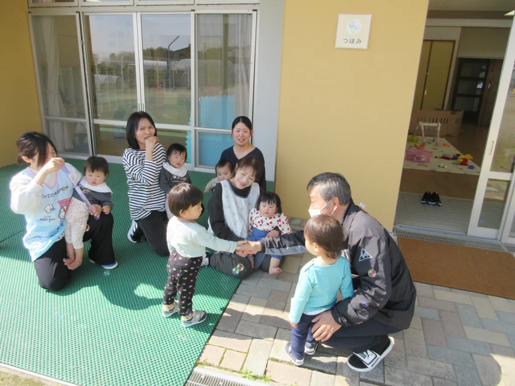 Caregivers and children sit and stand on a green outdoor mat outside a daycare, as a man in a mask shakes hands with a young child.
