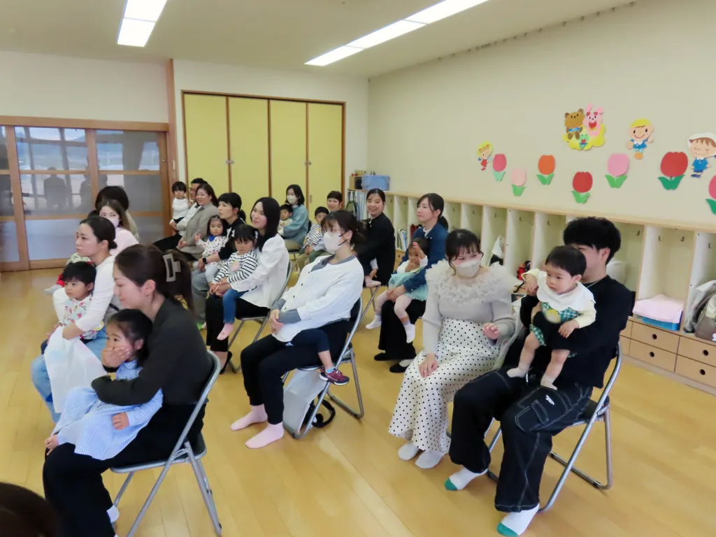 Adults and children sit on folding chairs in a bright classroom, watching a performance or presentation.