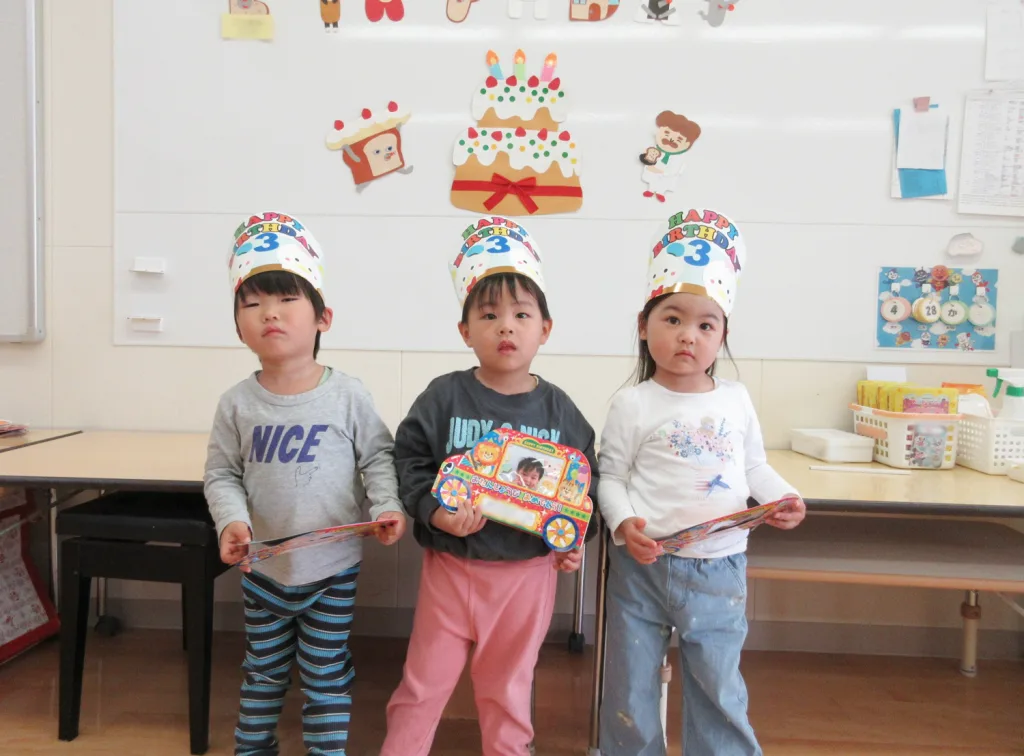 Three young children wearing birthday hats stand in a classroom, holding colorful party cards.
