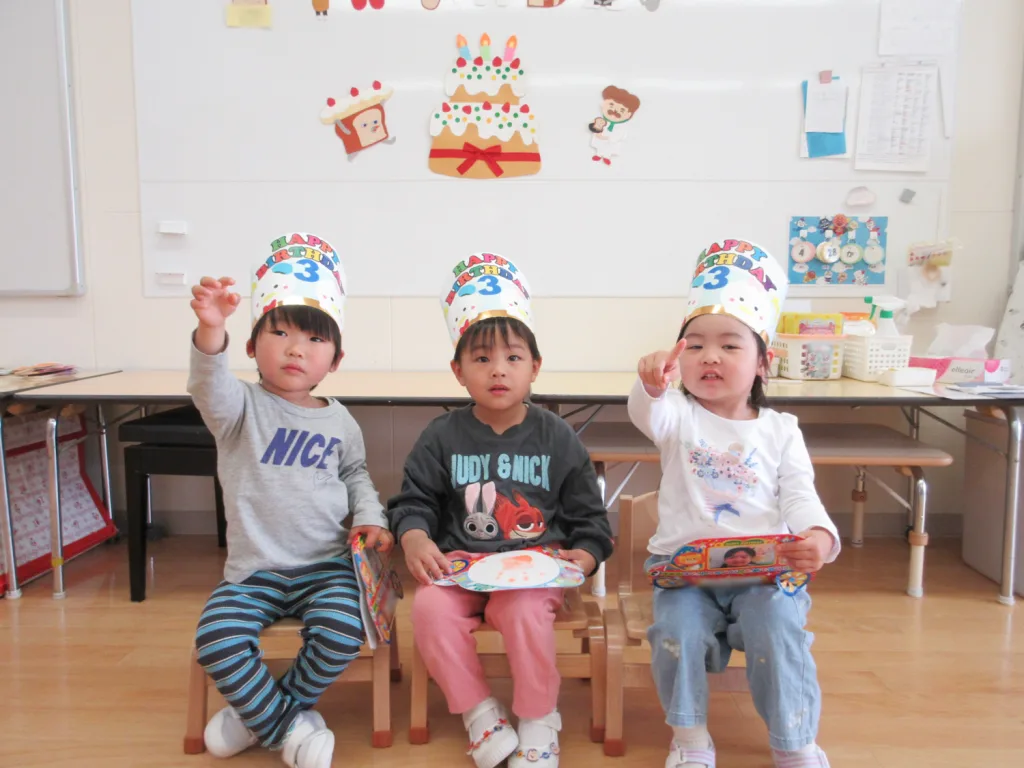 Three young children sit on small chairs in a classroom, wearing colorful birthday hats that read 'Happy Birthday 3' and raise their hands toward the camera.