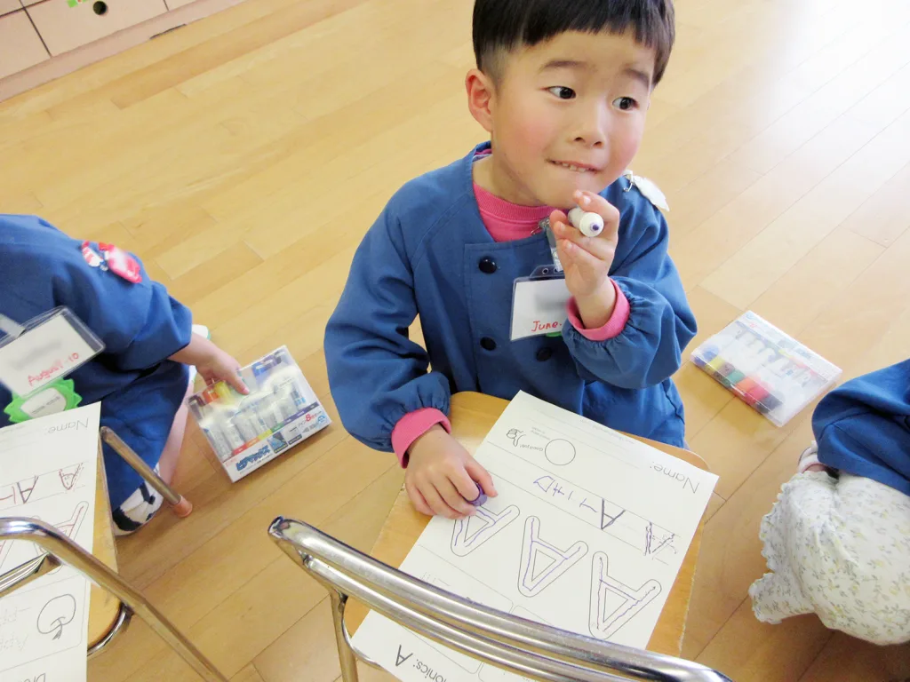 Young child in a blue smock holding a marker, looking to the side while working on a letter-writing worksheet at a wooden table with pencils and markers nearby.