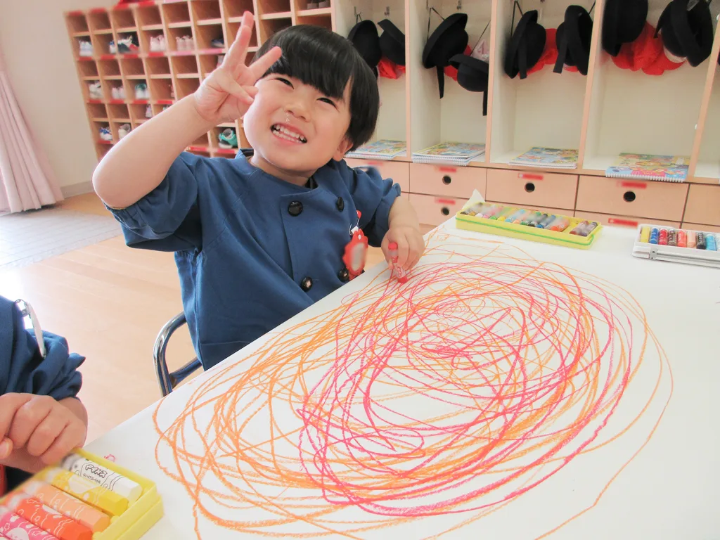 Young child in a blue smock smiles and makes a peace sign while drawing colorful scribbles with crayons at a classroom table, cubbies in the background.