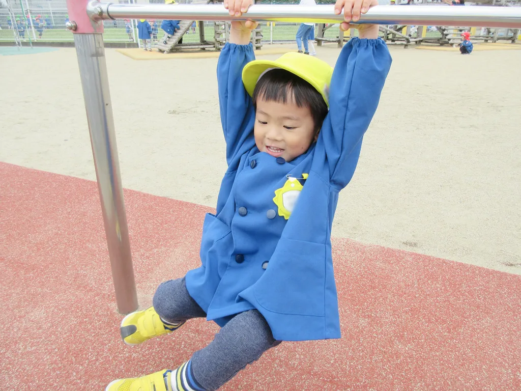 Smiling child in a blue coat and yellow cap hanging from a playground bar at a park.