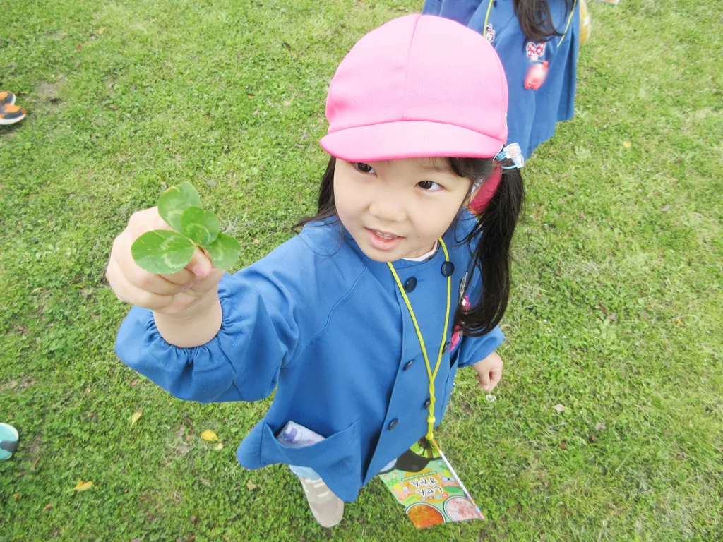 Young girl in a pink cap offering a four-leaf clover outdoors on grass field, wearing a blue jacket and a yellow lanyard.