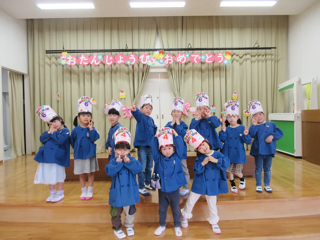Group of children in blue coats posing on a stage with birthday hats and a colorful 'Happy Birthday' banner above them.