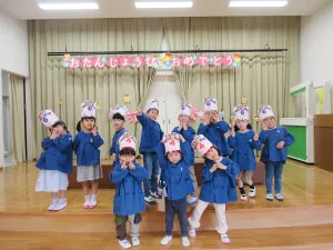 Group of children in blue coats posing on a stage with birthday hats and a colorful'Happy Birthday' banner above them.
