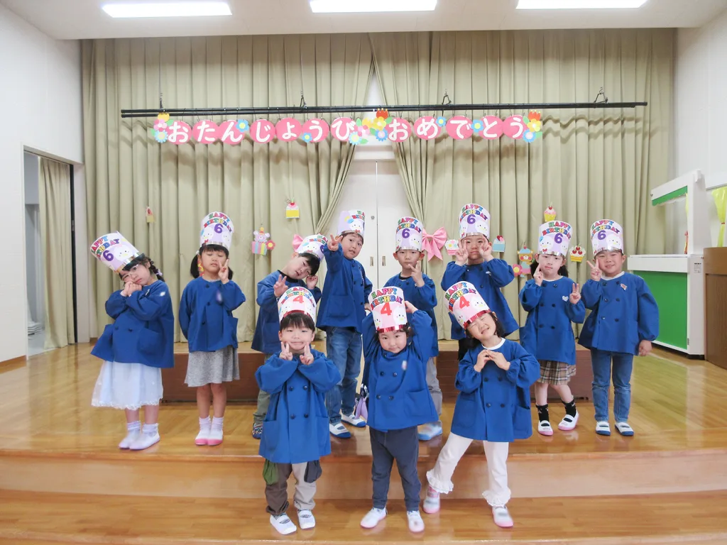 Group of happy children in blue coats on a stage, wearing birthday hats labeled 'Happy Birthday 6', posing with peace signs.