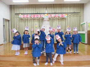 Group of happy children in blue coats on a stage, wearing birthday hats labeled'Happy Birthday 6', posing with peace signs.