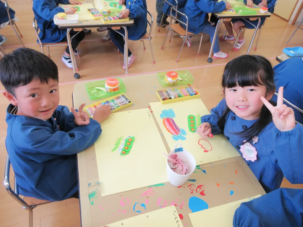 Two smiling children in blue smocks sit at a classroom table coloring with markers and paper art supplies on the table.