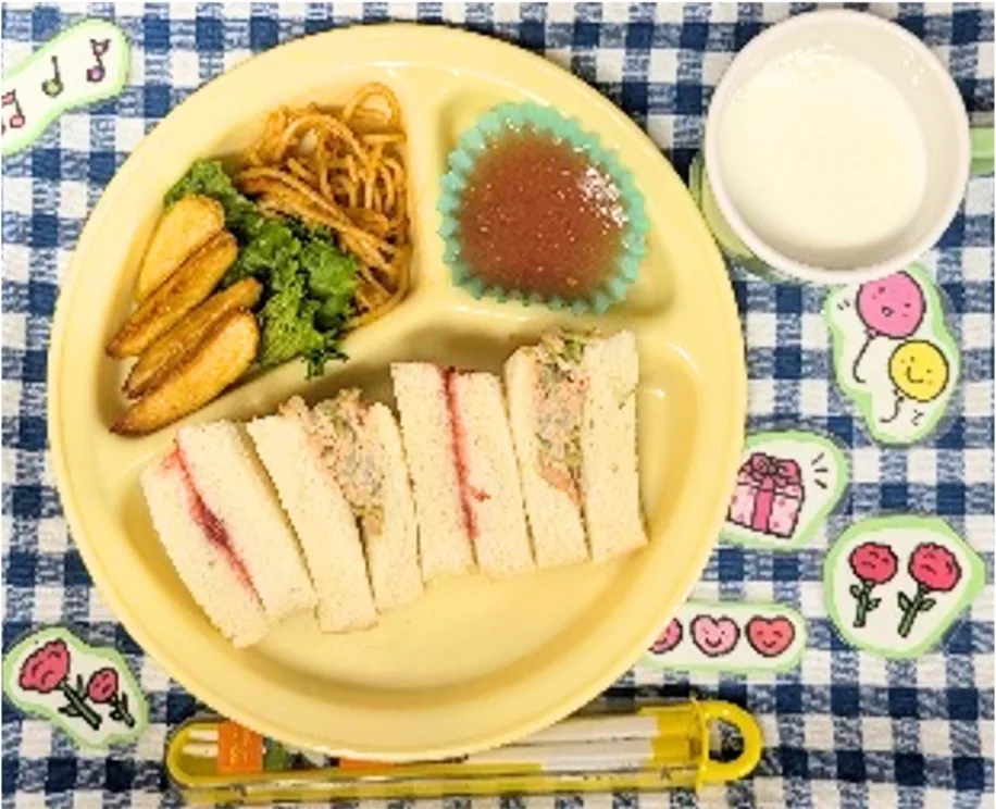 Yellow divided plate with finger sandwiches, potato wedges, greens, noodles, a jelly cup, and a cup of milk on a blue check tablecloth.