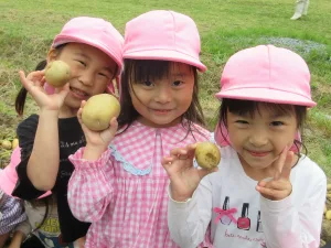 Three young girls wearing pink hats hold potatoes they harvested in a green field.