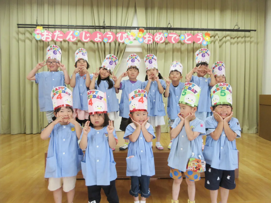 Group of children in light blue uniforms posing on a stage, wearing colorful birthday hats and flashing peace signs.