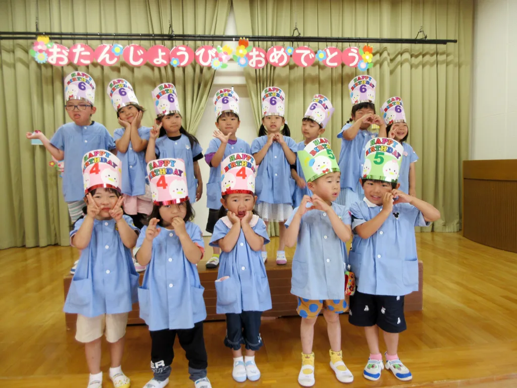 Group of children in blue uniforms posing on a stage, wearing colorful birthday hats and smiling.