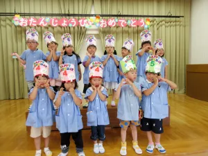 Group of children in blue uniforms posing on a stage, wearing colorful birthday hats and smiling.