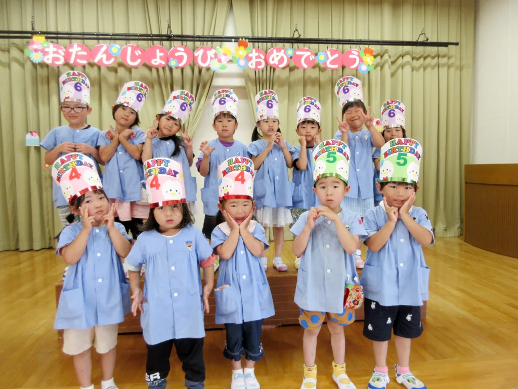 Group of children in blue outfits wearing birthday hats at a classroom birthday party, posing on a stage. 주요 포인트: 축하 분위기와 팀워크처럼 보이는 사진이네요.