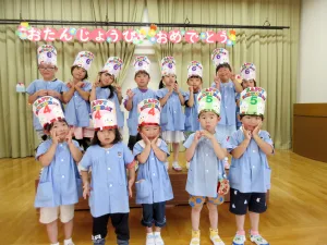 Group of children in blue outfits wearing birthday hats at a classroom birthday party, posing on a stage. 주요 포인트: 축하 분위기와 팀워크처럼 보이는 사진이네요.