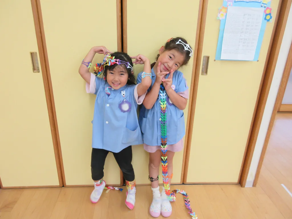 Two smiling girls in blue smocks pose by yellow lockers, wearing colorful paper chain necklaces and headbands.