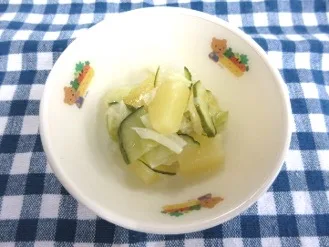 Small white bowl containing sliced cucumbers and pale yellow pickled vegetables on a blue-and-white checkered tablecloth, with decorative stickers around the rim.