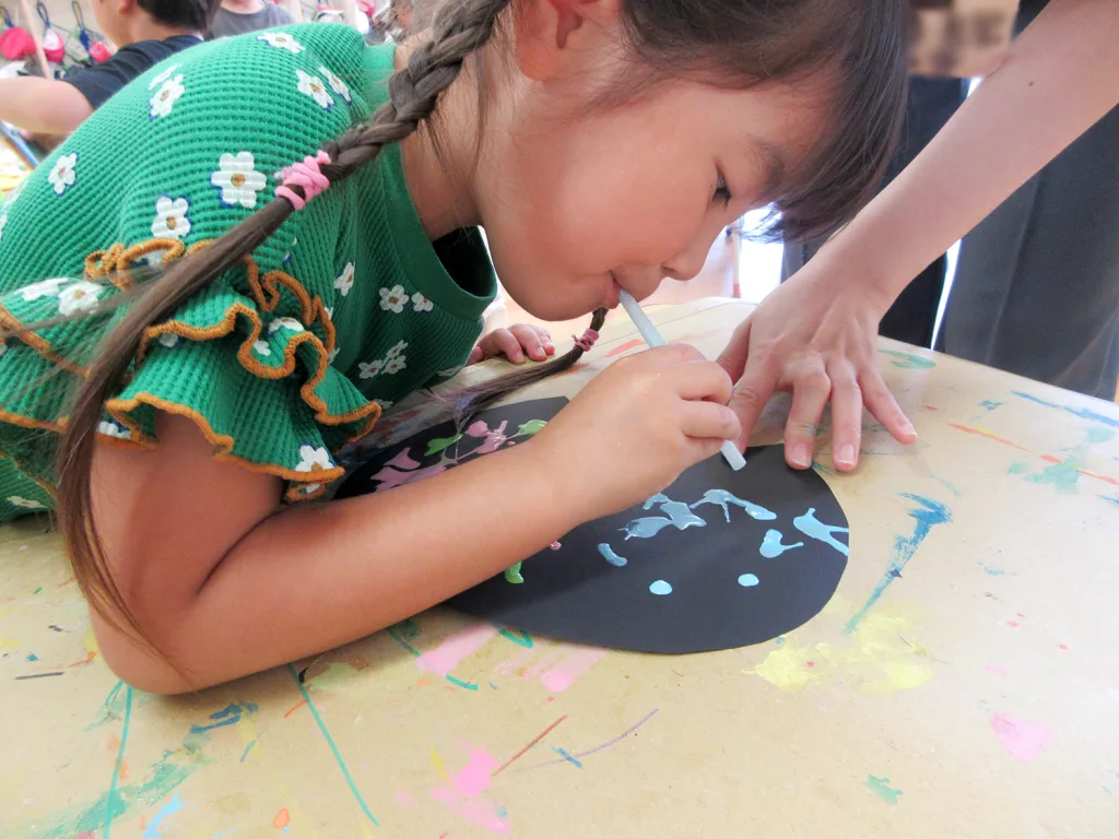 A young girl in a green floral sweater leans over a black circular paper, drawing with a light blue tool as an adult guides her hands during a colorful art activity.