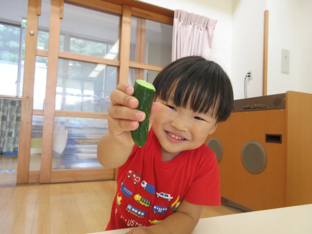 Smiling young child in a red shirt holds up a green cucumber toward the camera indoors, showing the cut end of the cucumber.