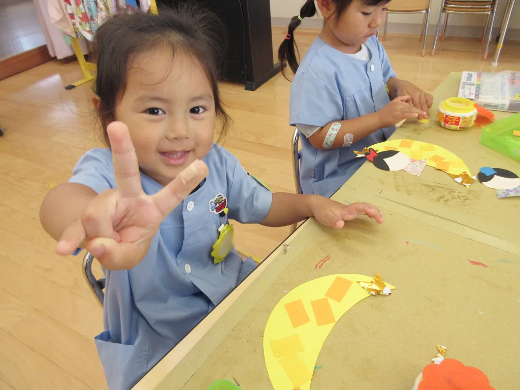 Two young children in blue smocks sit at a craft table, one girl showing a peace sign toward the camera while the other colors yellow paper pieces into a collage.