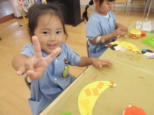 七夕飾りを作ったよ - 幼稚園型認定こども園 元岡幼稚園 - 幼稚園型認定こども園 元岡幼稚園 A smiling preschool girl in a blue smock holds up a peace sign toward the camera while crafting at a table with yellow paper mosaics.