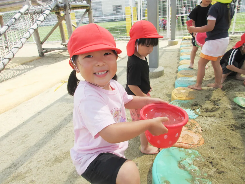 Smiling child in a pink shirt and red hat holds a red water bucket while playing in sandbox with other kids nearby.