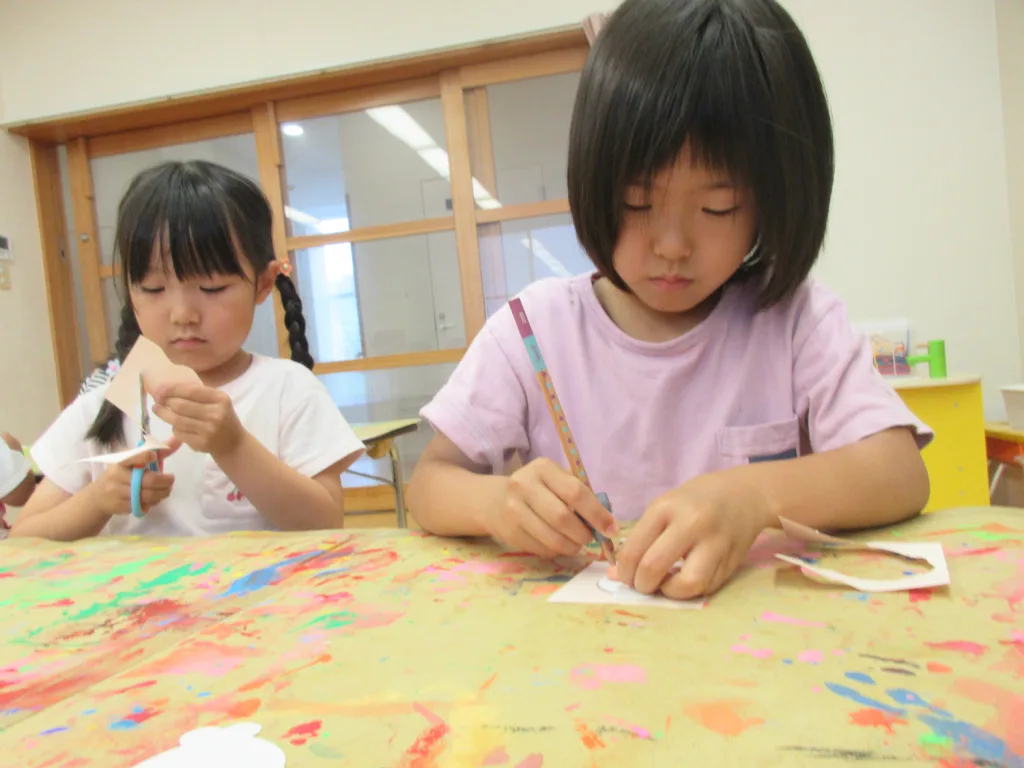 Two young girls sit at a colorful art table, engaged in crafts with scissors and pencils.