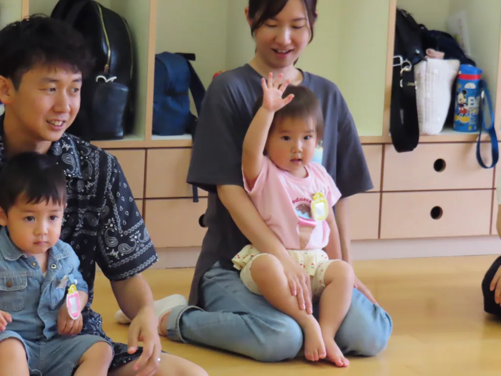 Caregiver sitting on the floor with a toddler on her lap, the child raises a hand during a group activity in a playroom.