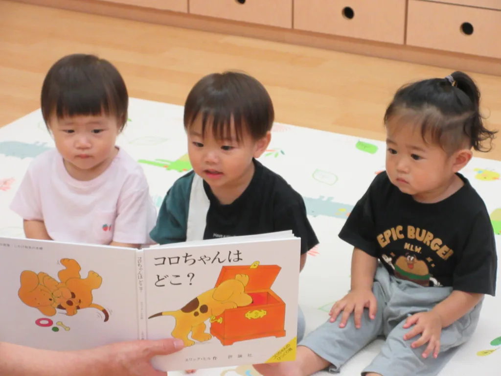 Three toddlers sit on a colorful play mat as an adult reads a Japanese children's book with cartoon dogs on the page.