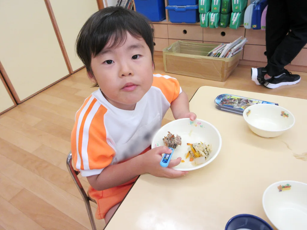 Child in an orange-and-white shirt sits at a classroom table, eating from a white bowl with a blue utensil, looking at the camera.