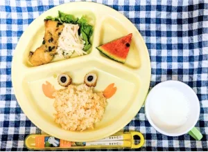 Divided yellow plate with crab-shaped rice, olive eyes, carrot claws, fried chicken with greens, a watermelon slice, and a cup of milk for a child lunch.