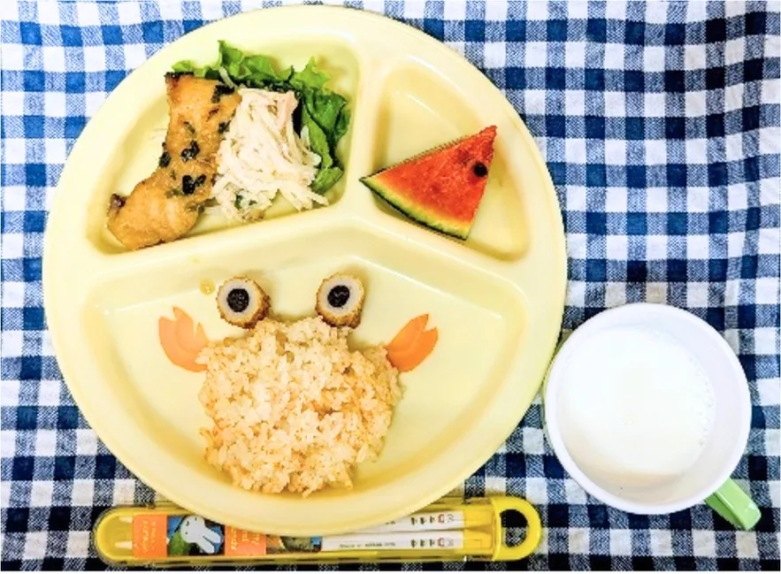 Divided yellow plate with crab-shaped rice, olive eyes, carrot claws, fried chicken with greens, a watermelon slice, and a cup of milk for a child lunch.