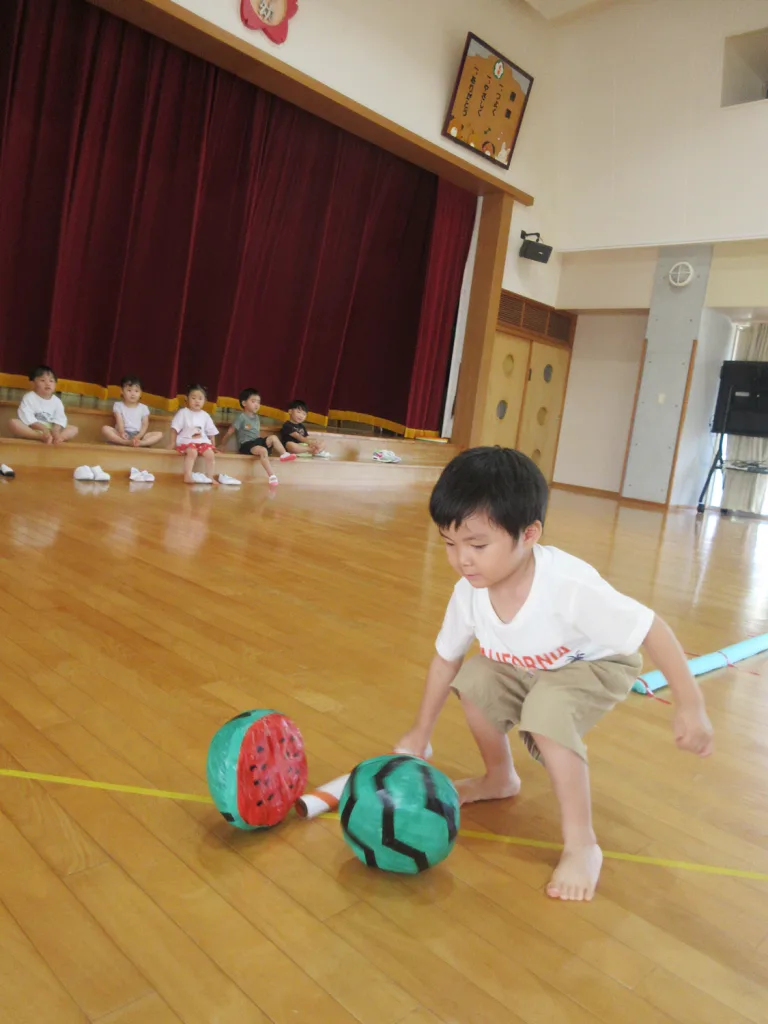 A young boy in a white shirt and tan shorts balances near two watermelon-patterned balls on a gym floor while other children sit along the stage in the background.