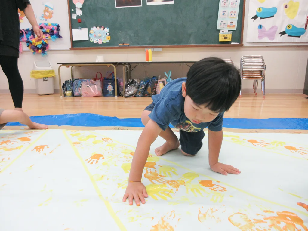 Young child crawling on a wide paint-covered mat, making handprints during a classroom art activity.