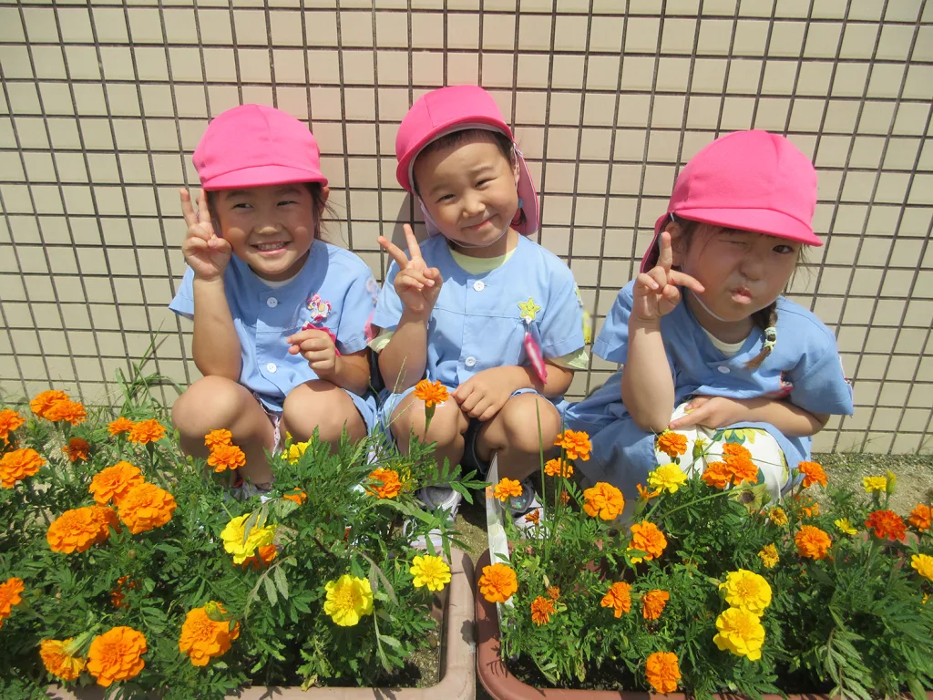 Three young girls in blue uniforms and pink hats sit by a flower bed, making peace signs at the camera against a tiled wall.
