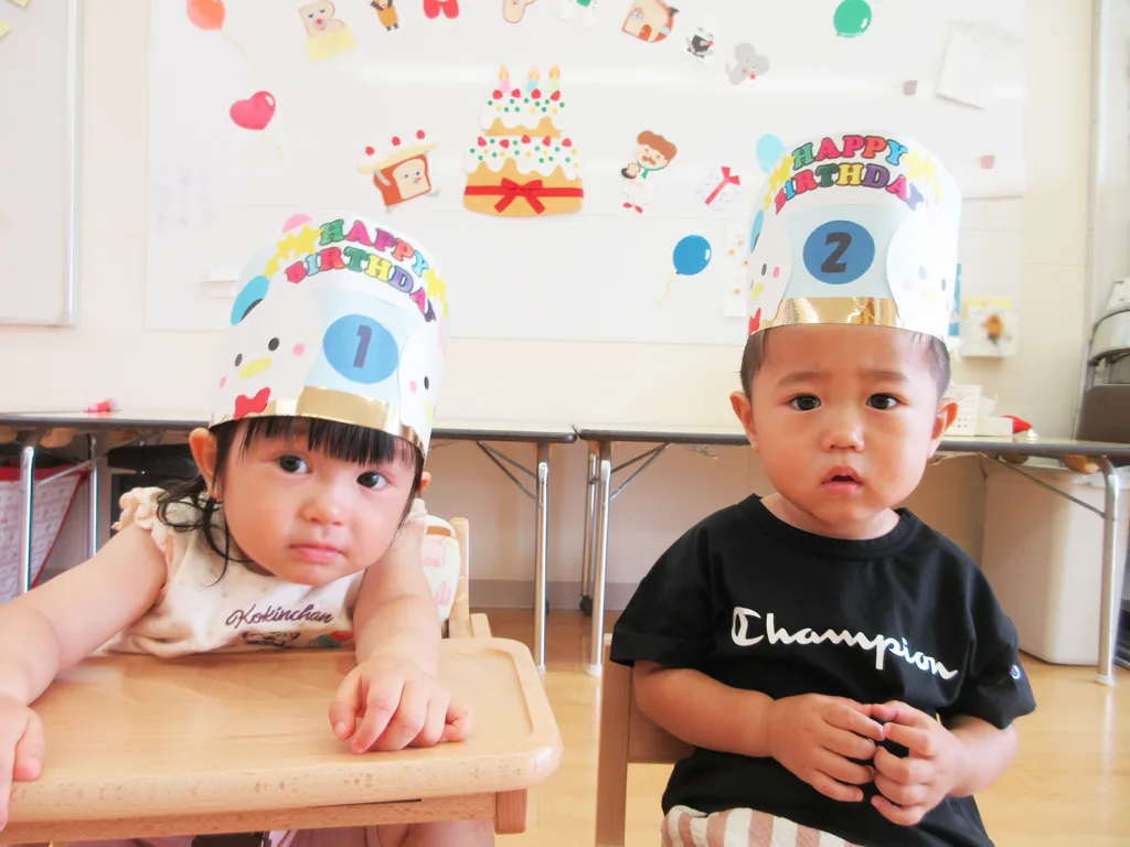Two young children wearing colorful birthday crowns that read 'Happy Birthday' with the numbers 1 and 2, seated at a wooden table in a bright classroom.