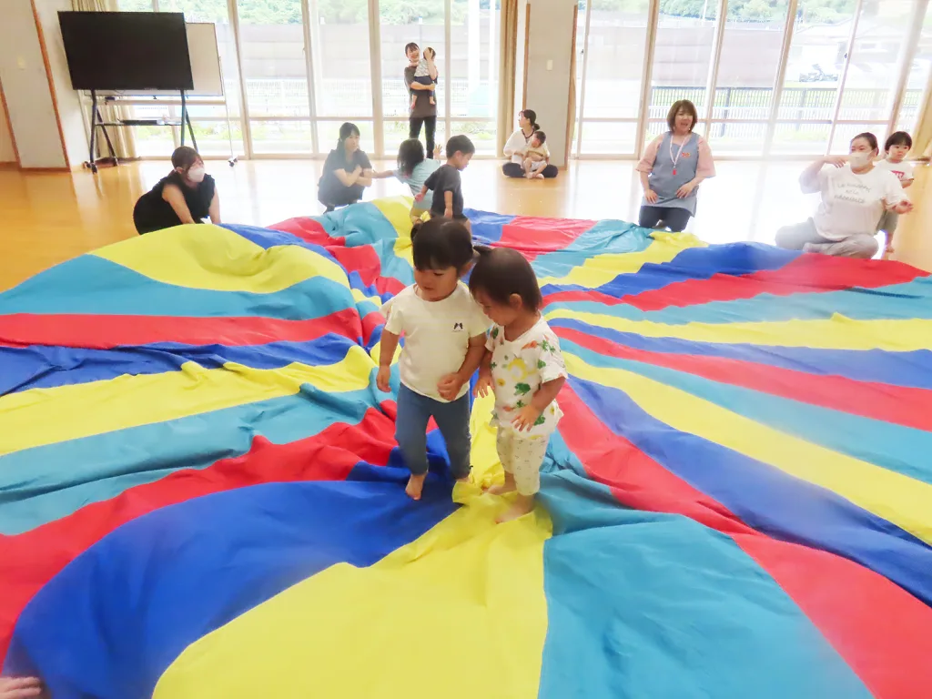 Two young children walk hand in hand across a large rainbow parachute laid on the floor while adults watch nearby in a bright room.