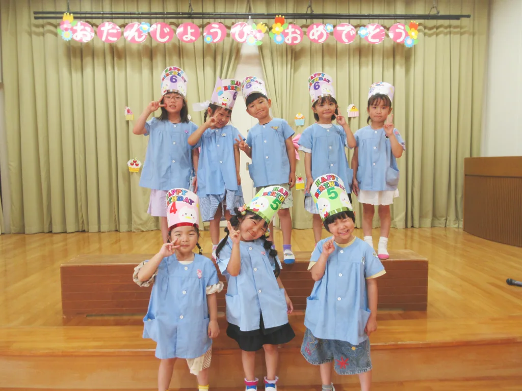 Group of children in blue uniforms posing on a stage with colorful birthday hats and a celebratory banner in the background.