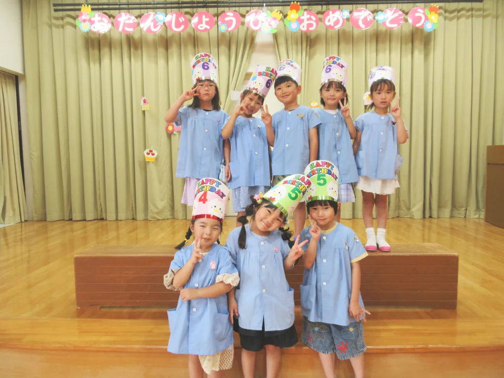 Group of birthday-clad children in blue uniforms posing on a stage with colorful 'Happy Birthday' hats and banner above.