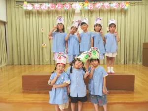 Group of birthday-clad children in blue uniforms posing on a stage with colorful'Happy Birthday' hats and banner above.