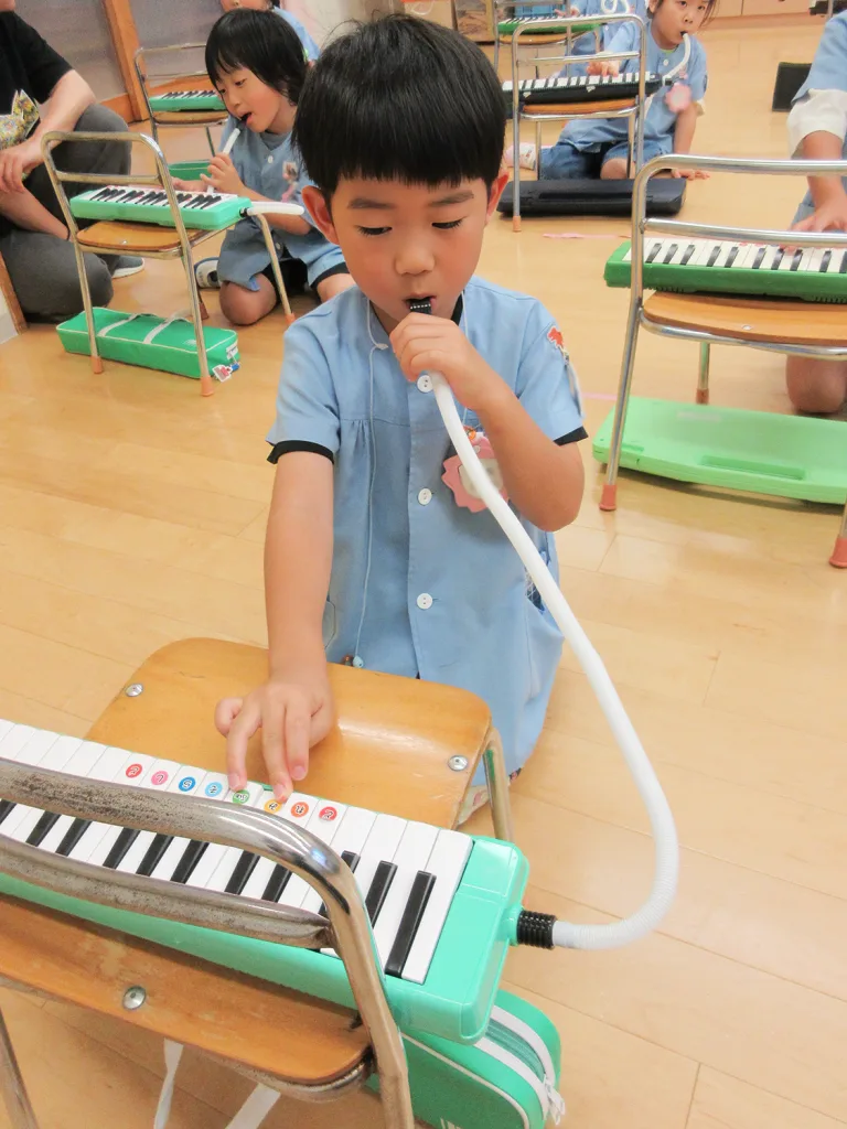 Boy in blue uniform plays a green melodica with a white mouthpiece in a classroom setting.
