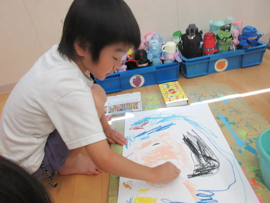 Young child kneels on the floor, drawing with crayons on a large sheet of paper with colorful scribbles.
