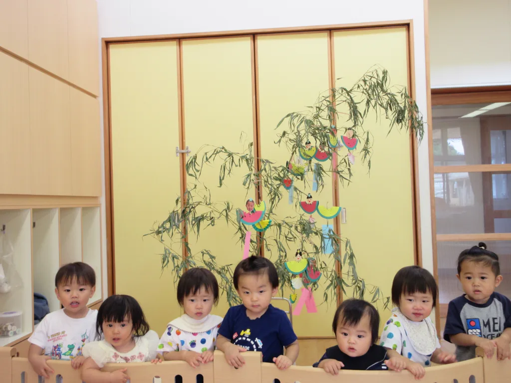 Group of toddlers seated at a table in a classroom with a decorated bamboo branch and colorful paper ornaments behind them, likely for Tanabata festival.