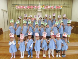 Group of young children in blue uniforms posing on a stage with birthday hats and a colorful banner that reads Happy Birthday in Japanese above them.