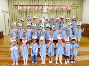 Group of young children in blue uniforms on a stage, wearing colorful birthday hats and posing for a birthday celebration photo.