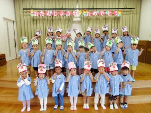 Group of preschoolers in blue uniforms posing on a stage for a birthday celebration, wearing colorful birthday hats and a'Happy Birthday' banner above them.