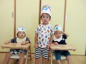 Three toddlers in birthday hats pose indoors: a standing child in the middle with striped shorts, flanked by two seated siblings in wooden high chairs.